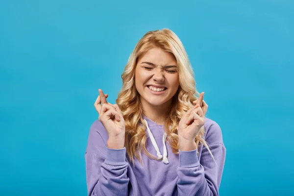 Tensa rubia adolescente con los ojos cerrados sosteniendo los dedos cruzados y deseando suerte en azul - foto de stock