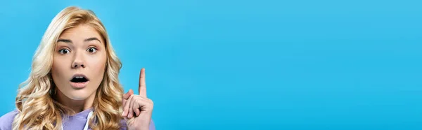 Animado loira adolescente com boca aberta mostrando sinal de ideia no azul e olhando para a câmera, banner — Fotografia de Stock