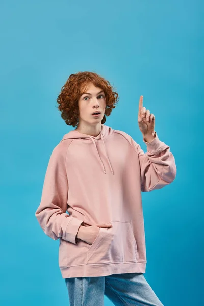 Amazed redhead teen guy showing idea sign, holding hand in pocket and looking at camera on blue — Stock Photo