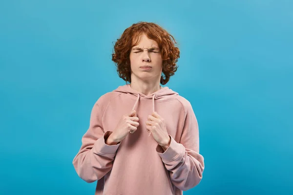 Nervous boy with red wavy hair wearing trendy hoodie and standing with closed eyes on blue — Stock Photo