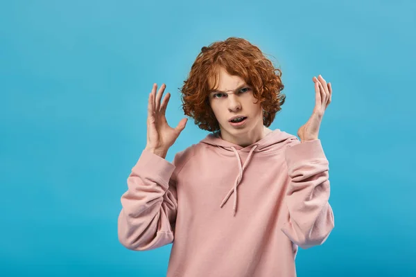 Раздраженный подросток с рыжими волнистыми волосами, жестикулирующий и смотрящий в камеру на синем, плохом настроении — стоковое фото