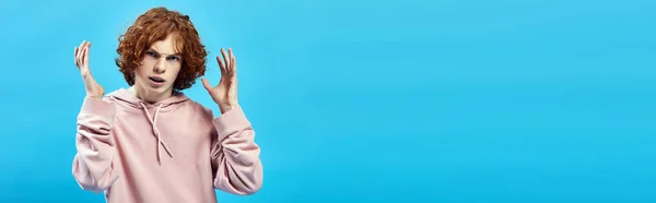 Angry teenage guy with red wavy hair looking at camera and gesturing on blue, bad mood, banner — Stock Photo