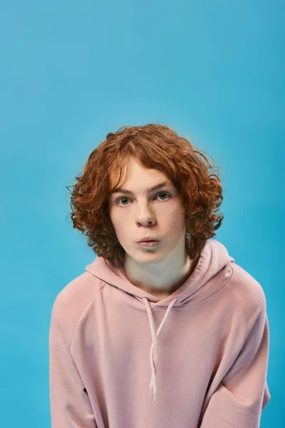 Portrait of impressed teenage boy with red wavy hair in stylish hoodie and looking at camera on blue — Stock Photo