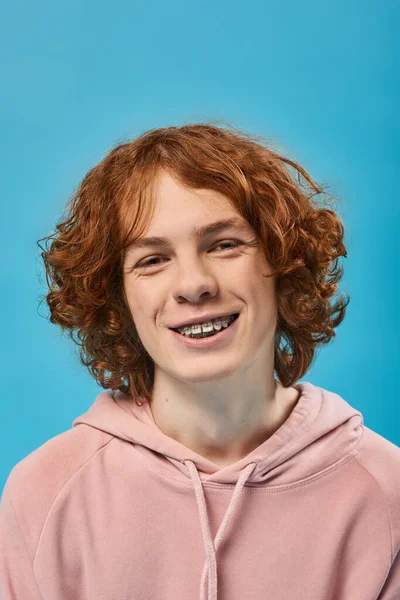 Portrait of cheerful teenage guy with red wavy hair and braces smiling at camera on blue, happiness — Stock Photo