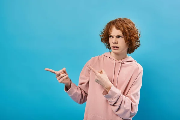 Disgustado pelirroja adolescente en sudadera con capucha de moda mirando hacia otro lado y señalando con los dedos en azul - foto de stock