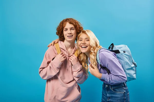 Estudiantes adolescentes alegres y elegantes con mochilas sonriendo a la cámara en azul, amigos felices - foto de stock