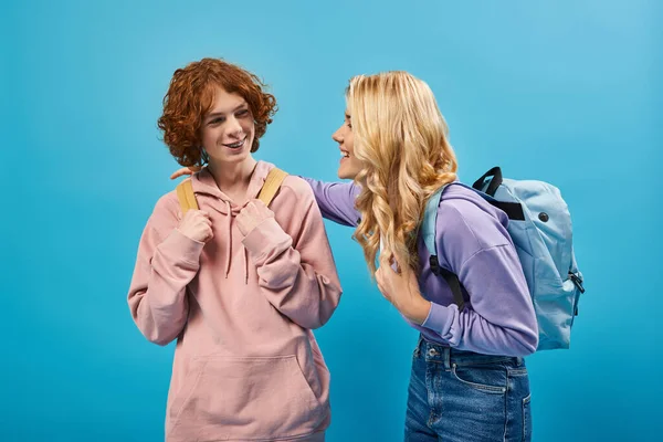 Rubia adolescente chica con escuela mochila abrazando sonriente pelirroja novio en azul, unidad y vinculación - foto de stock