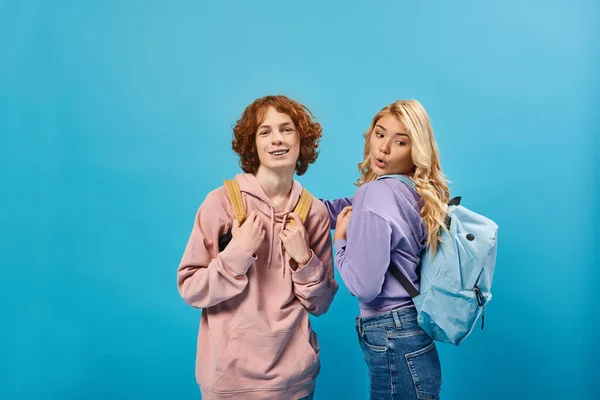 Изумленная блондинка-подросток смотрит на школьный рюкзак рядом с веселым рыжим одноклассником на синем — стоковое фото