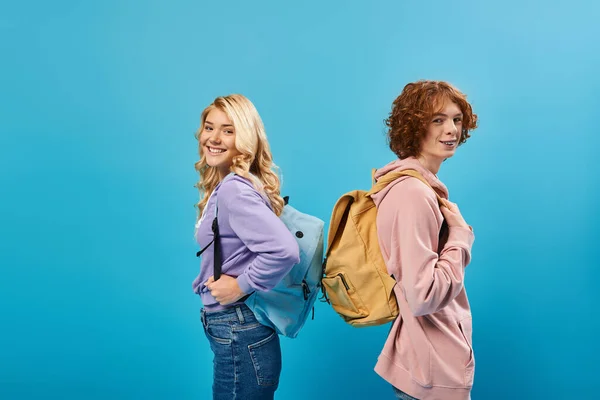 Feliz adolescente estudiantes en sudaderas con capucha de pie espalda con mochilas y mirando a la cámara en azul - foto de stock