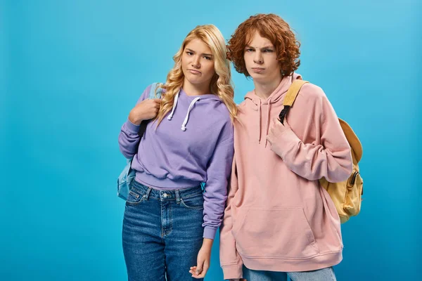 Displeased teenage classmates standing with school backpacks and looking at camera on blue — Stock Photo