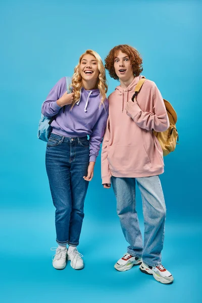 Cheerful teen students in trendy hoodies and denim jeans posing with backpacks on blue, full length — Stock Photo