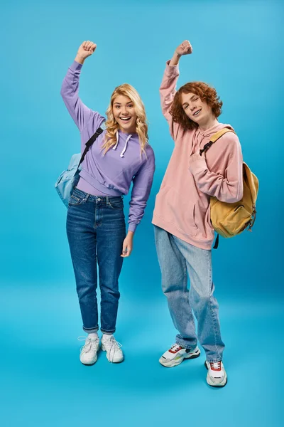 Estudantes adolescentes animados e na moda com mochilas mostrando gesto de vitória em azul, comprimento total — Fotografia de Stock