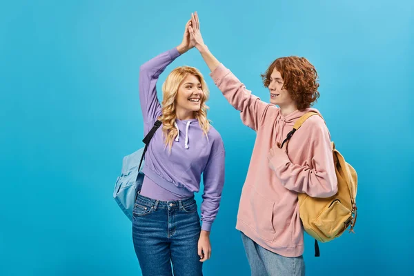 Colegas adolescentes felizes com mochilas vestindo roupas da moda e dando cinco no azul — Fotografia de Stock
