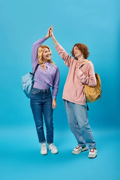 Estudiantes adolescentes de moda con mochilas que dan cinco altos mientras están de pie en azul, longitud completa - foto de stock