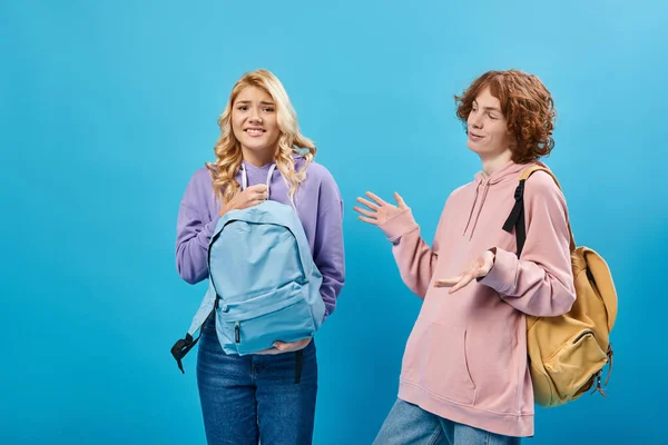 Cheerful redhead teen boy gesturing near displeased girlfriend holding heavy backpack on blue — Stock Photo