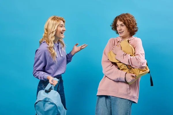 Laughing blonde teen girl talking to smiling redhead boyfriend holding school backpack on blue — Stock Photo