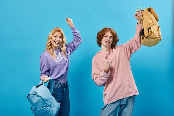 Excited teen students in trendy casual clothes holding backpacks and showing success gesture on blue — Stock Photo