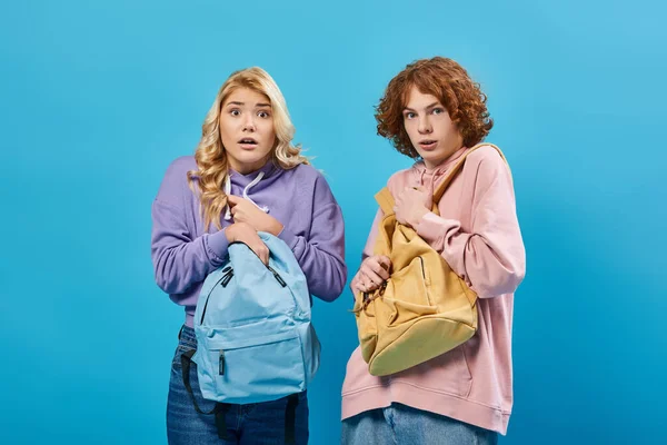 Испуганные и обеспокоенные подростки в толстовках, смотрящие в камеру и стоящие с рюкзаками на синем — стоковое фото