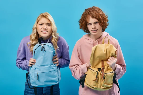 Estudiantes adolescentes enojados en sudaderas con capucha de moda sosteniendo mochilas escolares y mirando la cámara en azul - foto de stock