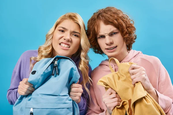 Angry and nervous teen students in with school backpacks grimacing and looking at camera on blue — Stock Photo