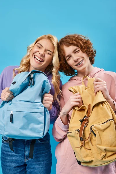 Emocionados y elegantes estudiantes adolescentes sosteniendo mochilas escolares y riendo con los ojos cerrados en azul - foto de stock