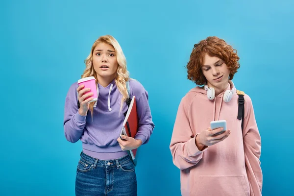 Besorgte blonde Teenagerin mit Pappbecher neben lächelndem Freund mit Smartphone und Kopfhörer auf blau — Stockfoto