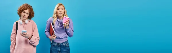 Усміхнений рудий хлопець використовує смартфон поруч стурбована дівчина з кавою, щоб піти блакитний, банер — стокове фото