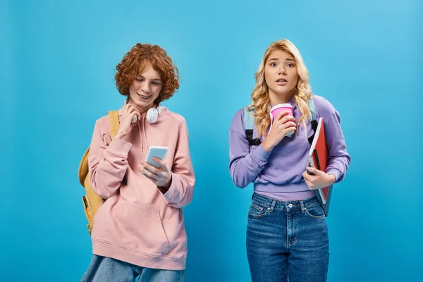 Нервова дівчина-підліток з паперовою чашкою дивиться на камеру біля позитивного друга, використовуючи смартфон на синьому — стокове фото