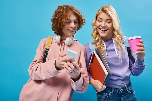 Pelirroja adolescente estudiante con auriculares mostrando smartphone a feliz novia con taza de papel en azul - foto de stock