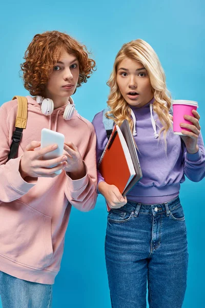 Amazed teenage students with backpacks, headphones and digital devices looking at camera on blue — Stock Photo