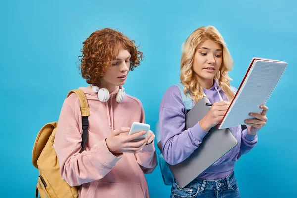 Девочка-подросток с ноутбуком, пишущая в блокноте рядом с рыжей подругой со смартфоном и наушниками на синем — стоковое фото