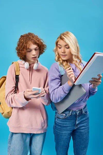 Rossa adolescente studente mostrando smartphone alla ragazza bionda con laptop e notebook su blu — Foto stock