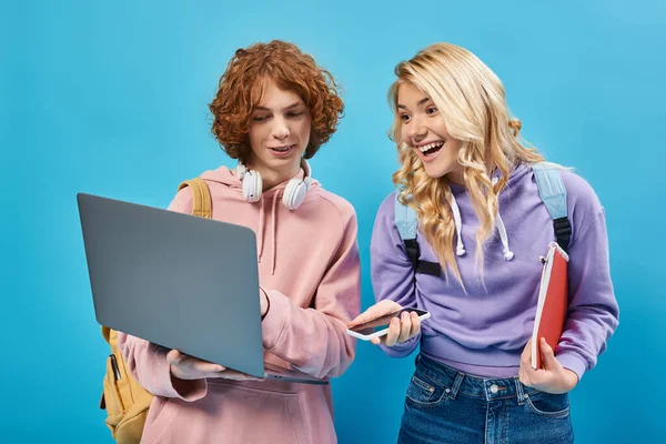 Excited teen girl with smartphone and notebook looking at laptop near redhead student on blue — Stock Photo