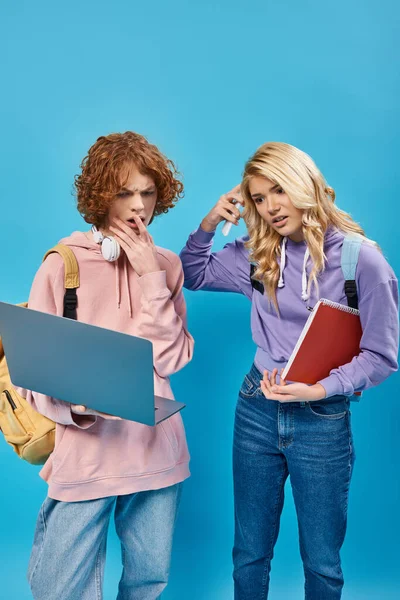 Shocked, thoughtful teen students with backpacks, headphones and notebook looking at laptop on blue — Stock Photo