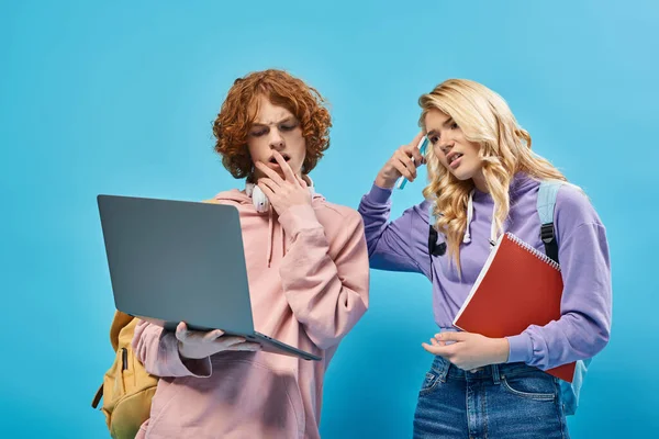Chocados y reflexivos compañeros de clase adolescentes con mochilas mirando a la computadora portátil en azul - foto de stock
