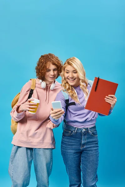 Excited teenage students with backpacks, notebook and coffee to go looking at smartphone on blue — Stock Photo