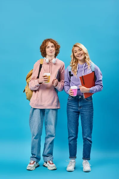 Cheerful teen students with backpacks and coffee to go in paper cups looking at camera on blue — Stock Photo