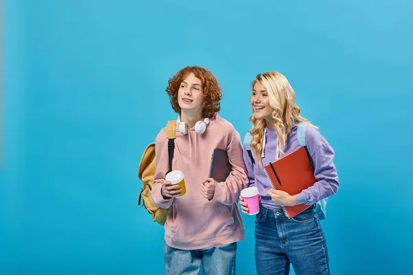 Estudiantes adolescentes alegres con mochilas y bebidas para llevar en vasos de papel mirando hacia otro lado en azul - foto de stock
