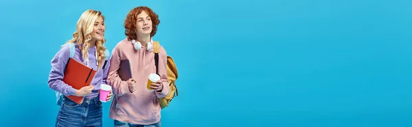 Happy teen students with backpacks and takeaway drinks smiling and looking away on blue, banner — Stock Photo