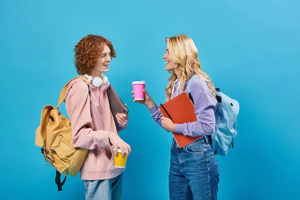 Happy blonde teen girl with notebook and paper cup talking to redhead student with laptop on blue — Stock Photo
