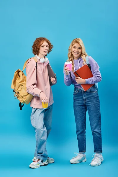 Cheerful and trendy teenage students with backpacks and coffee to go smiling at camera on blue — Stock Photo