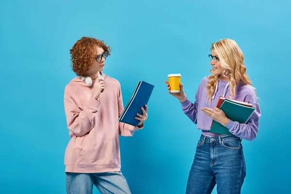 Teenage students in eyeglasses on blue holding notebooks and coffee to go while talking on blue — Stock Photo
