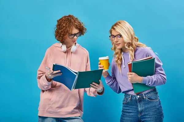 Вдумлива руда студентка-підліток показує блокнот блондинці подрузі з паперовою чашкою на синьому — стокове фото