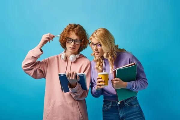 Thoughtful redhead student in eyeglasses holding notebooks near girlfriend with paper cup on blue — Stock Photo