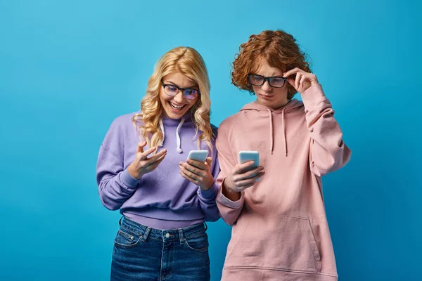 Cheerful blonde teen girl in eyeglasses looking at smartphone near thoughtful redhead friend on blue — Stock Photo