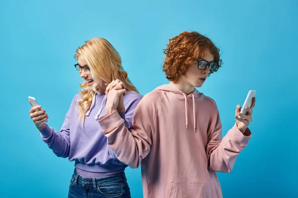 Teenage friends in eyeglasses and stylish hoodies using mobile phones and holding hands on blue — Stock Photo