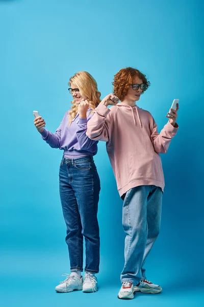 Happy and trendy teenage friends in eyeglasses and using smartphones and showing win gesture on blue — Stock Photo