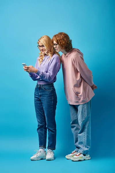Lächelndes Teenie-Mädchen in Brille und stylischer Kleidung mit Smartphone in der Nähe eines rothaarigen Freundes auf blauem Grund — Stockfoto