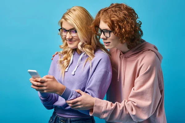 Joyful blonde teen girl in eyeglasses and trendy hoodie using smartphone near redhead friend on blue — Stock Photo