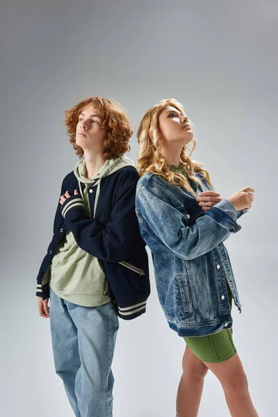 Dreamy teenagers in trendy casual attire standing back to back and looking away on grey — Stock Photo